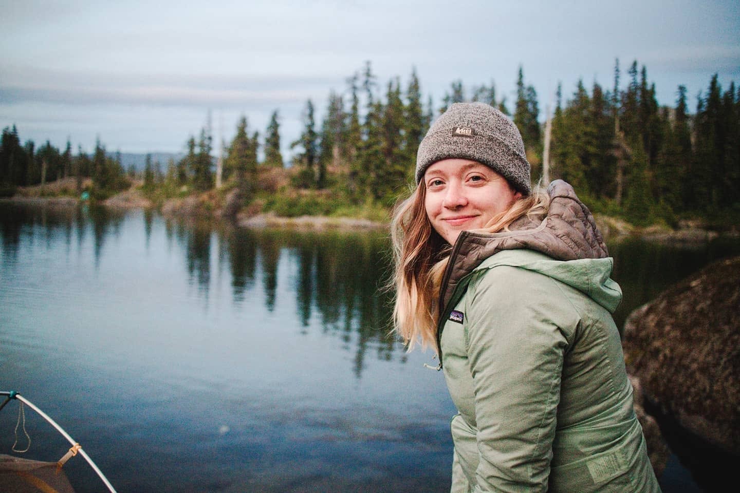 Chelsea Newton smiling over her shoulder beside a mountain lake, wearing a grey beanie and a green jacket