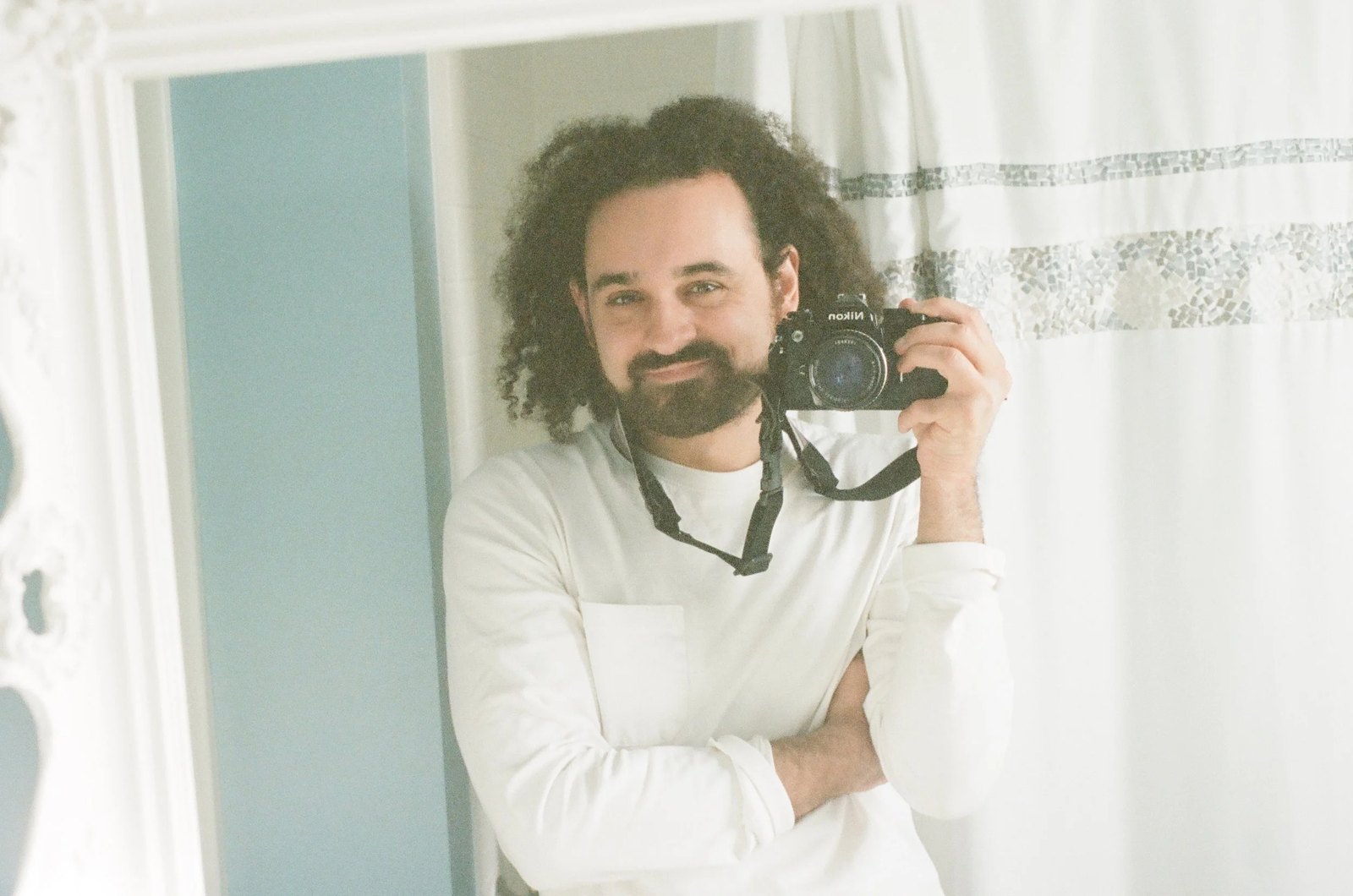 Jordan Bianchi smiling and holding a Nikon film camera, photographed in a mirror